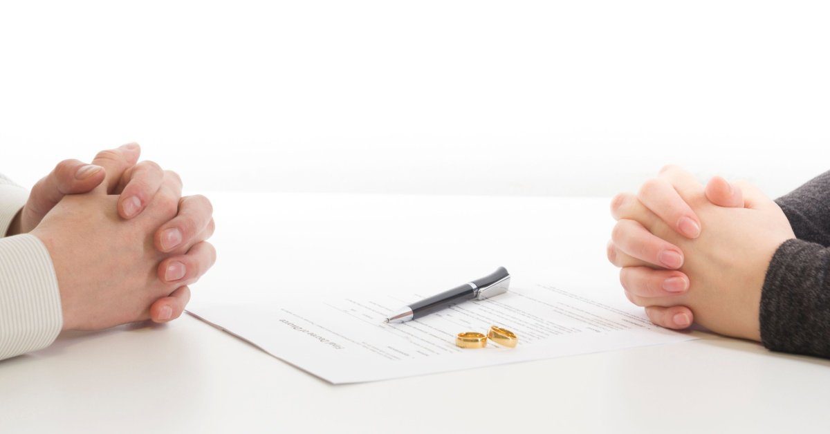 2 hands, fingers crossed, pen, paper and wedding rings on table, concept of Who Should File for Divorce First in NJ?